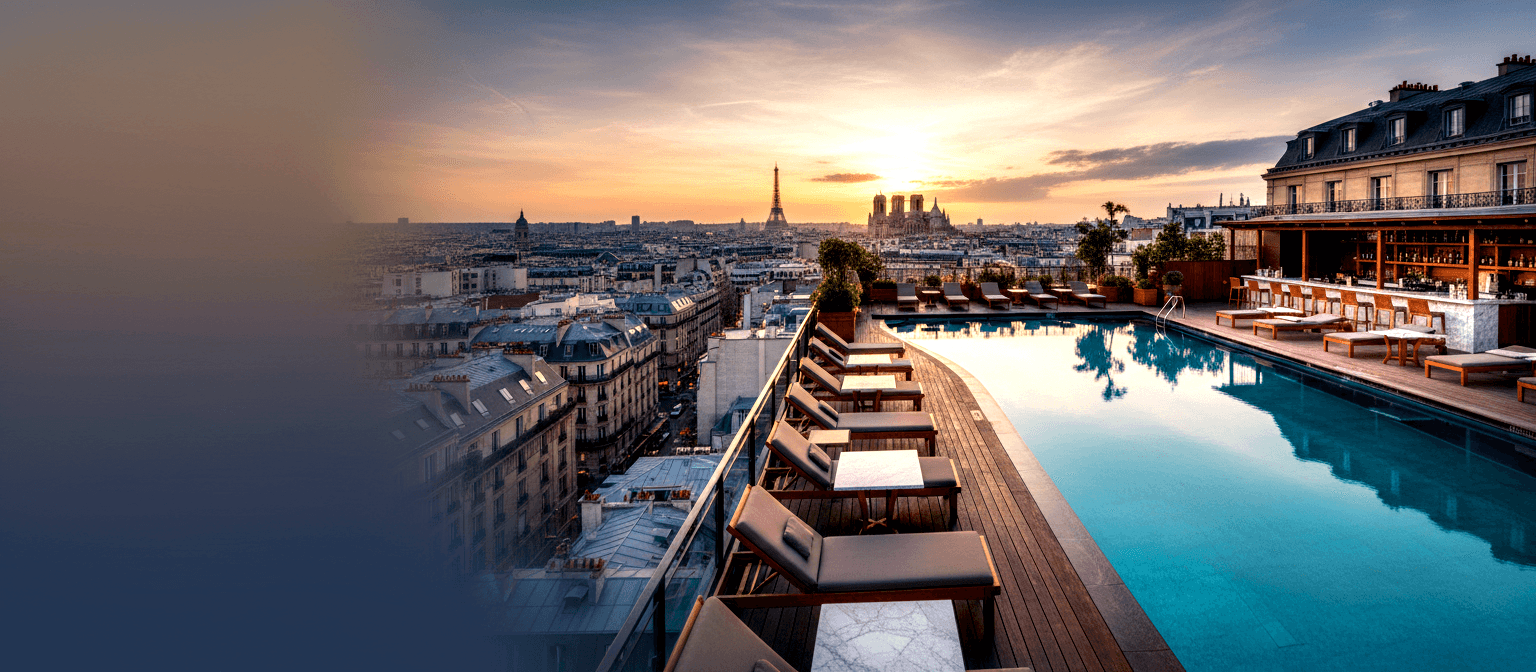 Rooftop infinity pool, glass balustrade, wooden loungers, covered bar; classical urban skyline under golden-orange sunset.