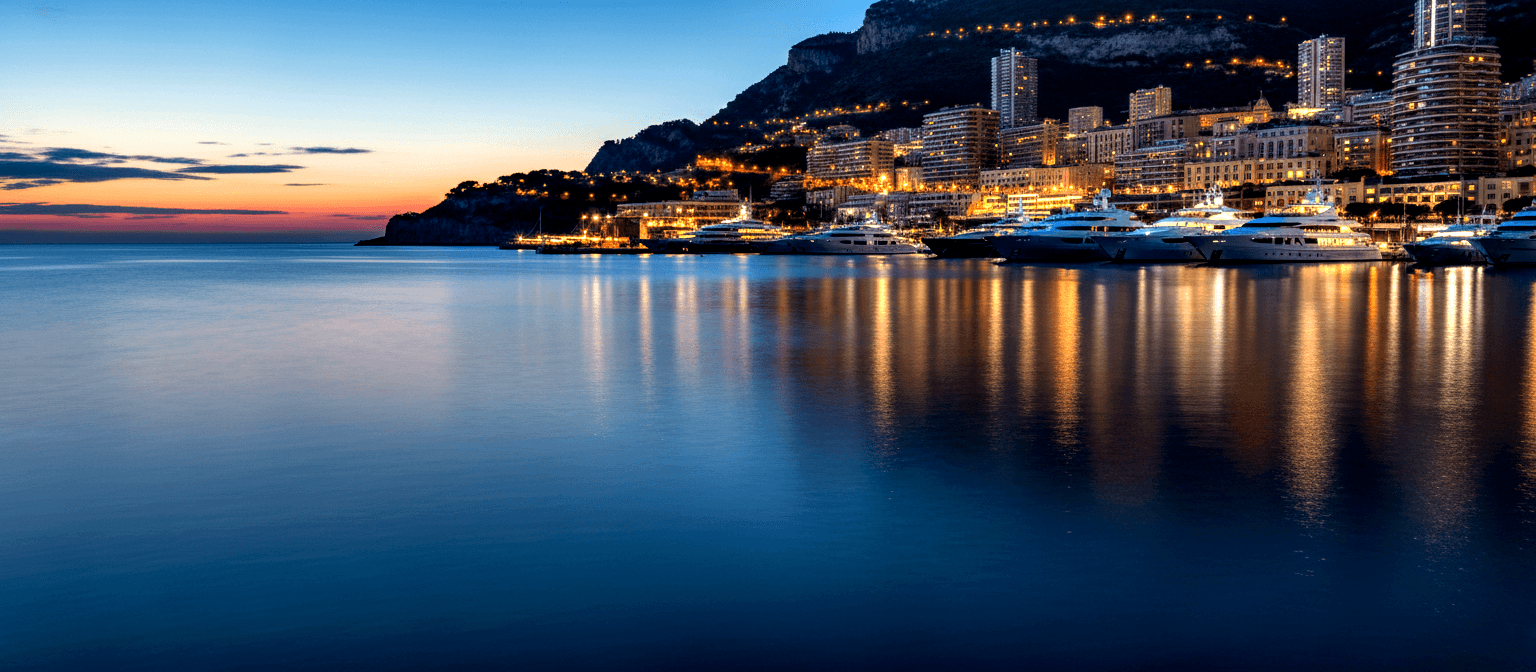 Harbor at blue hour: superyachts moored at marina, city buildings on rocky hillside, amber lights reflected in calm water.