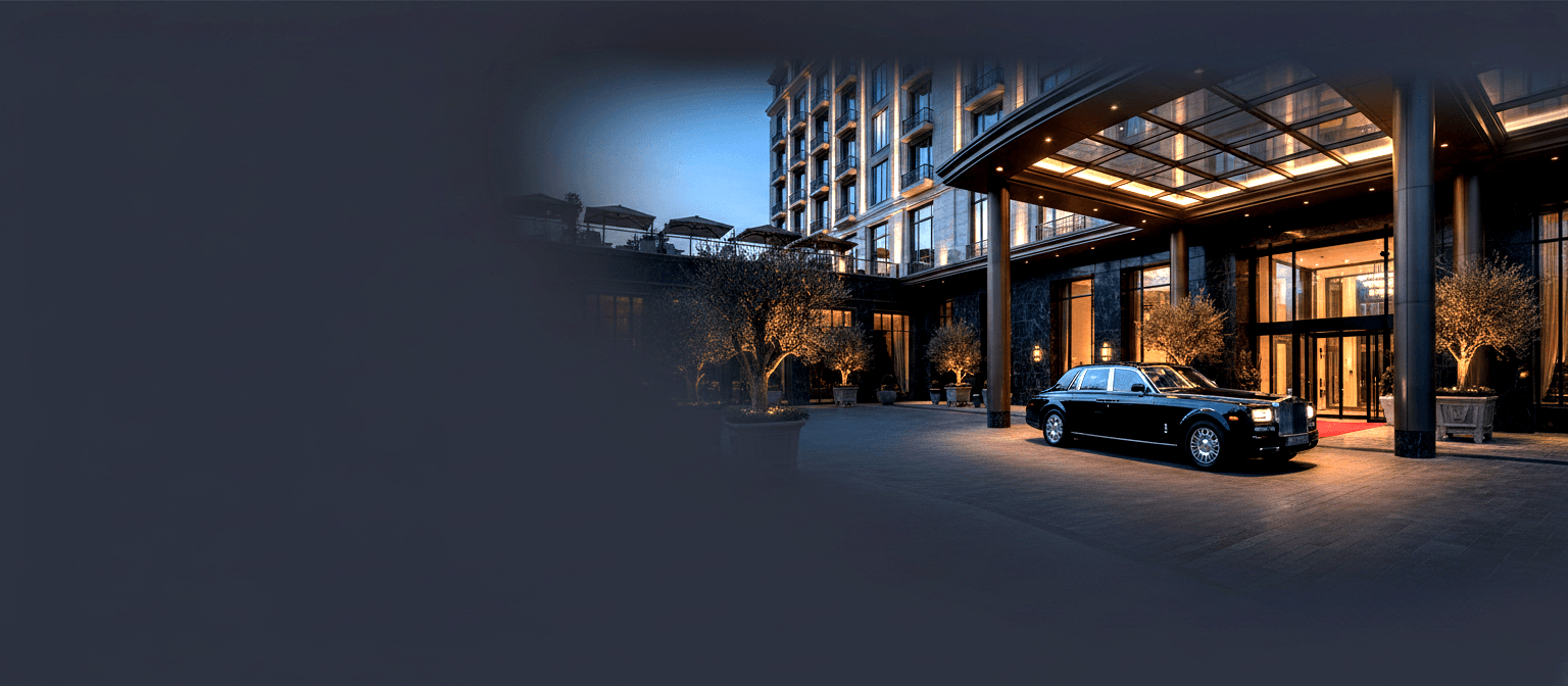 Black luxury sedan parked under a glass-and-steel porte-cochère at dusk, red carpet and revolving doors of a classical stone-façade building.